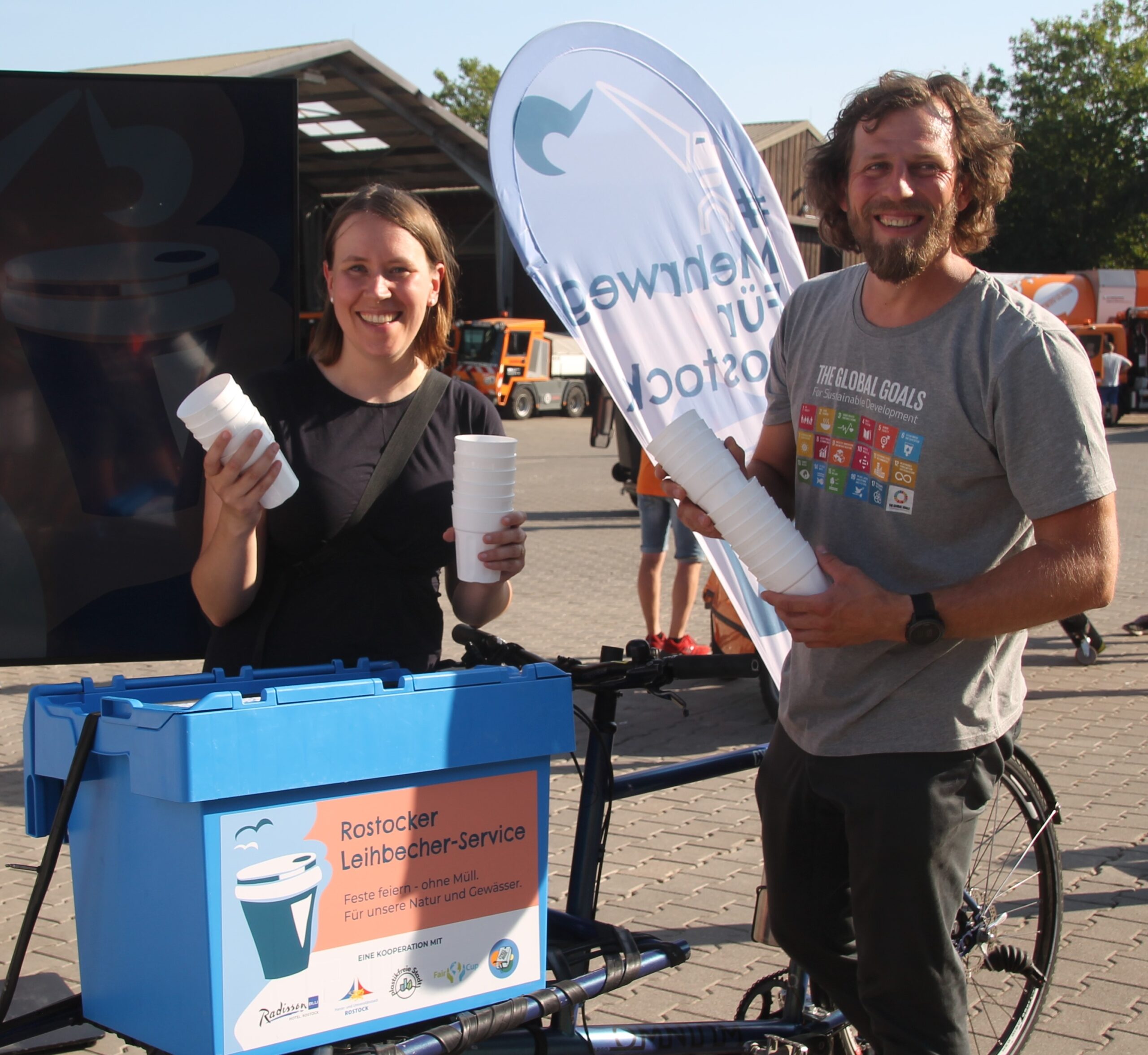 2 Mitarbeitende von Plastikfreie Stadt präsentieren eine Kiste mit Mehrwegbechern auf einem Lastenrad.